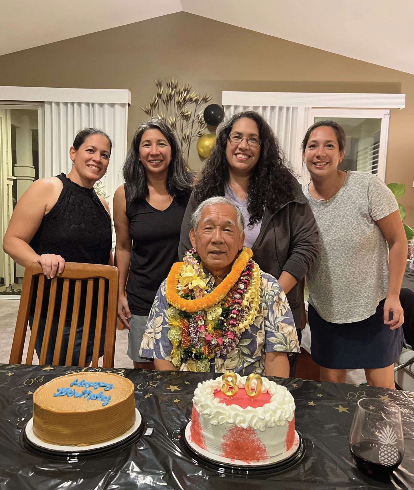 Shimoda and 3 other women smile and stand behind an older man wearing many leis who is seated in front of two birthday cakes